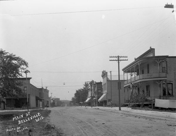 View down street of intersection at Main Street. The commercial street houses a bank, printing press, clothing shop, and hotel. A man makes repairs from a ladder at the Park Hotel.