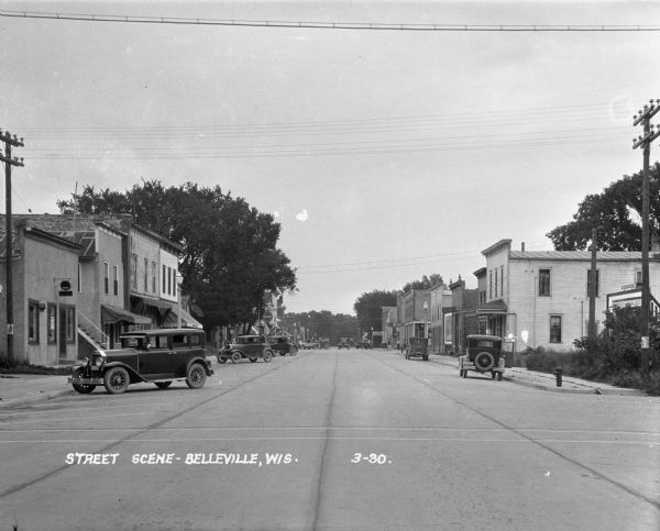 View looking down a commercial street. Shops and cars in angled parking spaces line both sides of the paved street. A horse-drawn wagon is in the far distance near a railroad crossing.