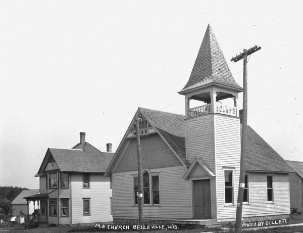 Exterior of the Methodist Evangelical Church. The building features a gable roof, a bell tower, a steeple, and double front doors. On the left is a house and behind it there appears to be a barn.