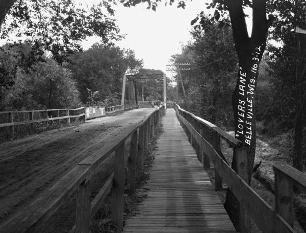 A bridge with both a wooden walkway for pedestrians and a dirt road for automobiles. Two men and a horse are standing on the other side of the bridge.