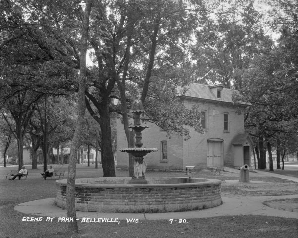 Water fountain near city hall at Library Park. There is a water fountain or bubbler on the edge of the fountain. Two men are sitting on separate benches in the background on the right.
