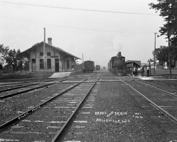 View down railroad tracks of the Belleville train depot. A group of people on the platform approach the train.