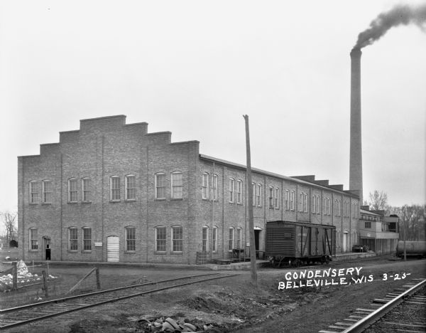 View across railroad tracks of a condensery building. The building features two-stories, a crow-stepping roof, and a tall chimney exhausting smoke. A railroad boxcar is parked alongside the building.