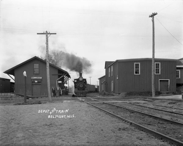 View down side of railroad tracks of locomotive pulling into the Belmont train station. A group of children stands at the platform.