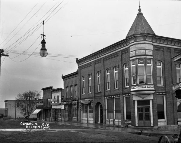 View across unpaved road of storefront on Commercial Avenue after the rain. There is a butcher shop, F. Tallada Candy and Cigars, and a saloon. A streetlight hangs from a utility line.