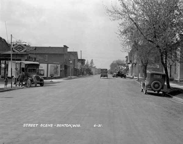 View down middle of street. On the left a man and two children stand near a truck parked in the driveway of a service station.
