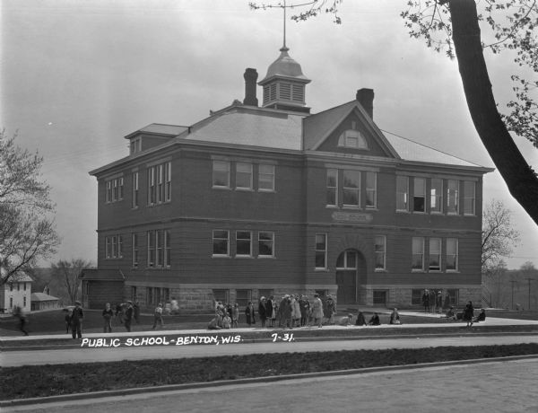 View from across street of schoolchildren standing and playing on the sidewalk outside the Benton High School.