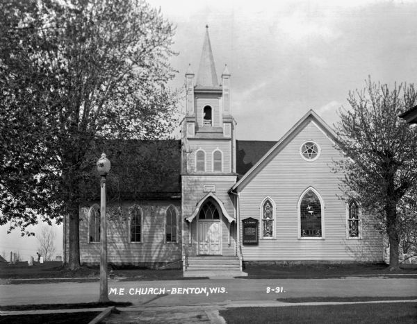 Exterior view from across street of the church. It has a bell steeple, stained glass windows, double french doors with an archway, and a wall gable facade. In the background a man is working in the cemetery which surrounds the church. The message board reads:   Sunday  9:30 - church school  10:30 - morning worship  7:00 - evening worship  Midweek meeting  7:00 - Thursday evening   [???] are welcome.