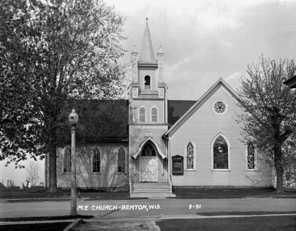 Exterior view from across street of the church. It has a bell steeple, stained glass windows, double french doors with an archway, and a wall gable facade. In the background a man is working in the cemetery which surrounds the church. The message board reads: 
 Sunday
 9:30 - church school
 10:30 - morning worship
 7:00 - evening worship
 Midweek meeting
 7:00 - Thursday evening 
 [???] are welcome.