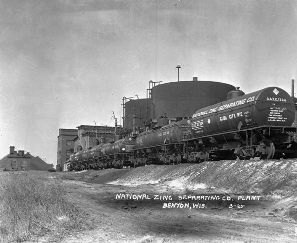 Railroad tank cars on railroad tracks outside the plant. There are two large round storage tanks behind the tank cars.