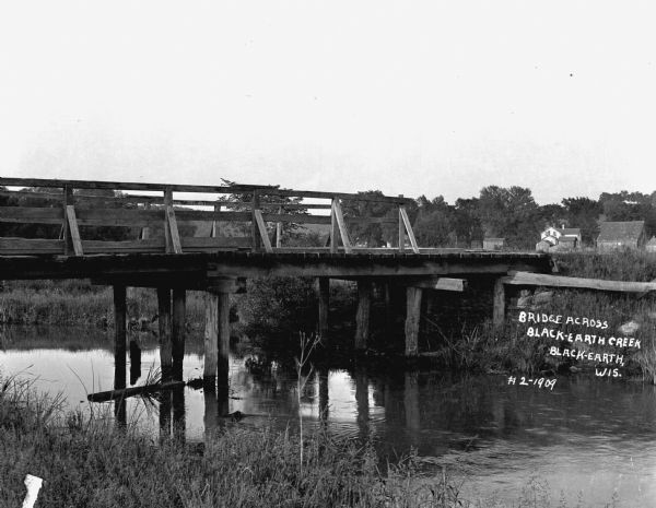View from shoreline of wooden bridge. There are homes and barns in the distance.