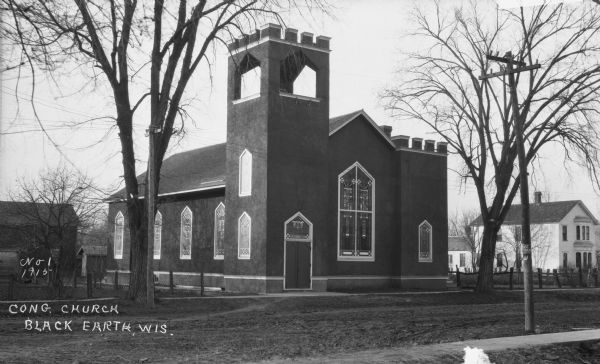 Exterior view of the church. The building features a bell steeple, stained glass windows, and double front doors. There is a fenced lot and a barn to the left of the church and a home on the right.