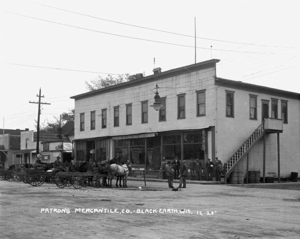 View from street of the Patrons' Mercantile Company consumer cooperative. A group of men stand outside the building. A row of horses and carriages are parked near the entrance along a railing.