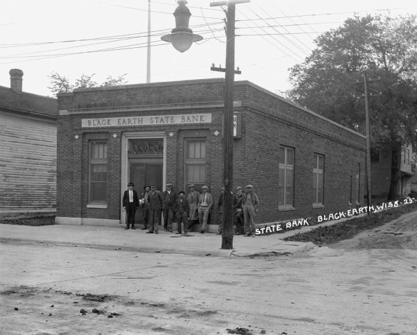 View across street of a group of men standing on the sidewalk in front of the bank. A streetlamp hangs from a utility line.