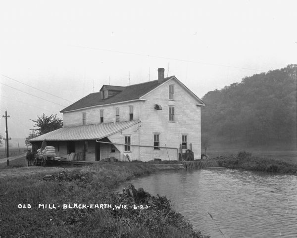 View along shoreline of a mill on a river. There is a man loading bags onto a cart at the front. There is a wooded hill on the right.
