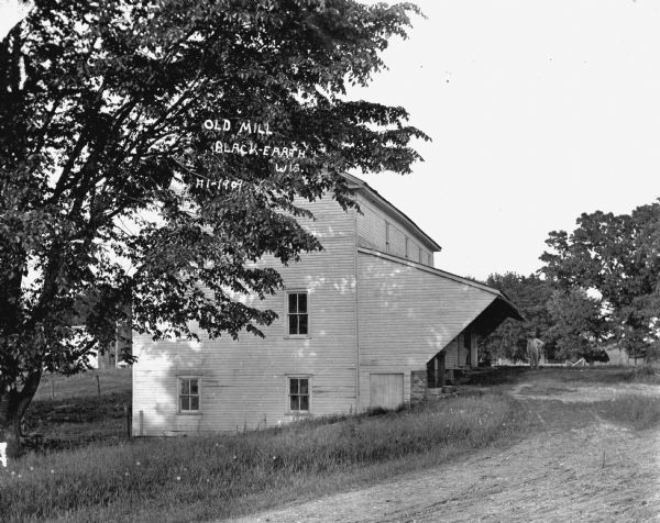 View of the side of a mill. The mill has a large awning over a loading dock. A man with a beard and a hat stands near the side of the mill.