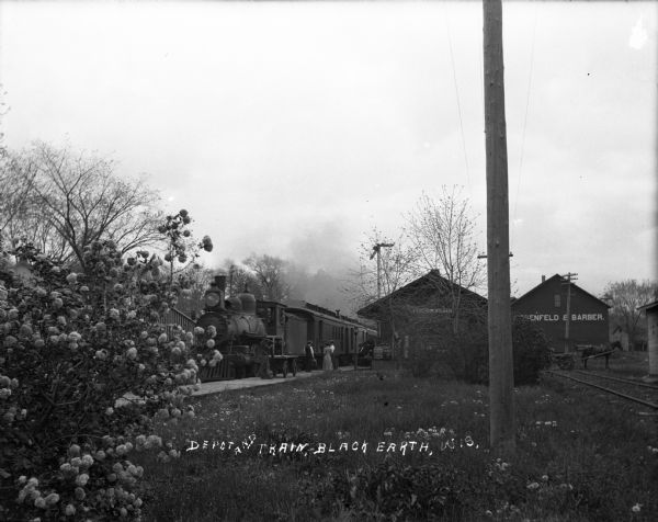 View from a distance of the train depot. On the left a locomotive is parked along the train platform where a group of people are standing. In the center is the depot partially obstructed by trees. On the right, there is a horse and cart, railroad tracks, and a building with a sign reading "Fesenfeld & Barber," a local hardware store.
