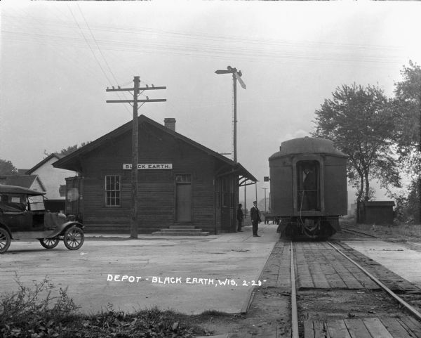 View of the train depot. A train is stopped at the depot. An automobile is parked on the left. A train conductor and a man stand at the outside.
