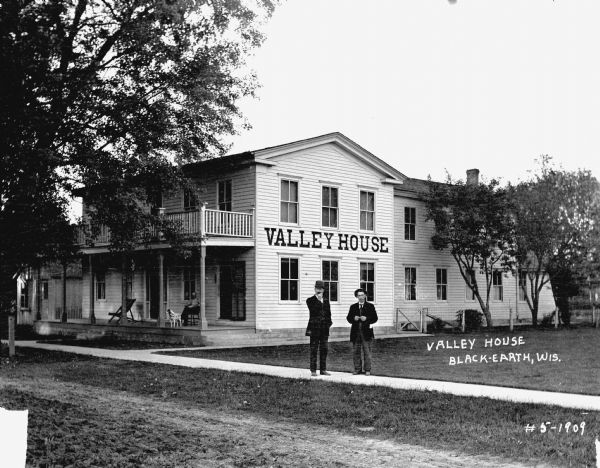 Two men stand on the sidewalk in front of a boardinghouse [?]. A man and a dog sit on the porch of the Valley House.