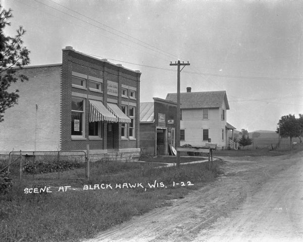 View from road of the Black Hawk State Bank, an automotive repair station, and a house. The Bank has striped awnings and a La Follette poster in the window. The automotive repair shop advertises Ford parts, Mobiloils, and Globe batteries. There are fields and hills in the distance.