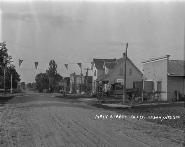 View down dirt road of right side of Main Street. Banners strung across the street read "Chautauga." There is a pickup truck parked in the automotive repair shop and gas station.