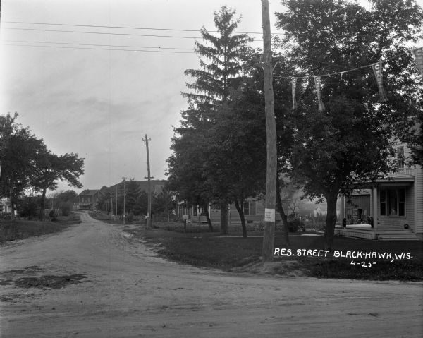 View down a winding dirt road with houses on both sides, as well as a large two-story building with a loading dock in front. Two people are in the back yard on the right near an outbuilding. There is a tree-covered ridge in the background.