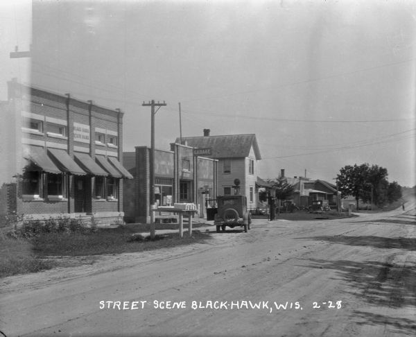View of the Black Hawk State Bank, an automotive repair and gas station and other shops. There is a car with its door open in front of the repair shop, near a group of mailboxes set up on a platform near the road. In the distance, a person stands in the road.