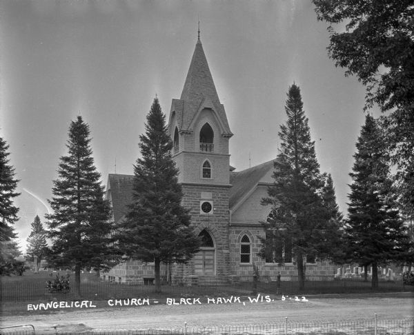 Exterior view from road of church. The church building features arched doors and windows, stained glass, and a bell steeple. There is a cemetery behind the church.