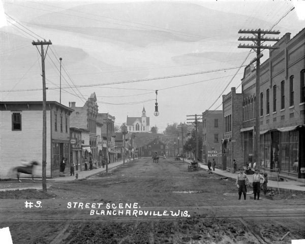 View down street of an intersection with railroad tracks and a streetlamp hanging from wires. There are storefronts on both sides of the street, and numerous pedestrians on the sidewalk. The visible shop signs are: S.M. Smith Eagle Saloon — billiard and pool upstairs, Bakery and Lunches, Barber Shop, T.S. Ryan Saloon, Hotel Ryan, Hotel Blanchardville, and Sherwin-Williams Paints. At the end of the street, there is a barn [?], and behind it on a hill is a church with a clock tower steeple. The time displayed is 7:40 [?].