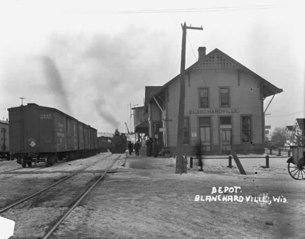 Exterior view of the Blanchardville Depot. The two-story building is painted with the village's name. A locomotive is in front of the platform where a group of people is standing. To the left are parked railroad cars.