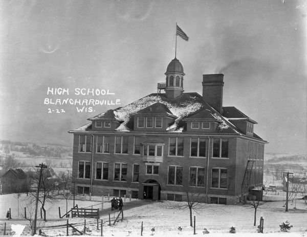 View of the high school. The building features an arched doorway, a hip roof with three dormers on the front and one on the side, a domed steeple with an American flag and widow's walk, a wide chimney, and a fire escape. On the walkway leading to the school, two men pose holding up a double bass. There is a playground with a swing-set to the right of the school.