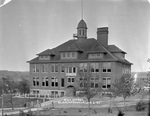 Elevated view of the high school. The building features an arched doorway, a hip roof with three dormers on the front and one on the side, a domed steeple with an American flag and widow's walk, a wide chimney, and a fire escape on the side. There is a playground with a swing-set to the right of the school.
