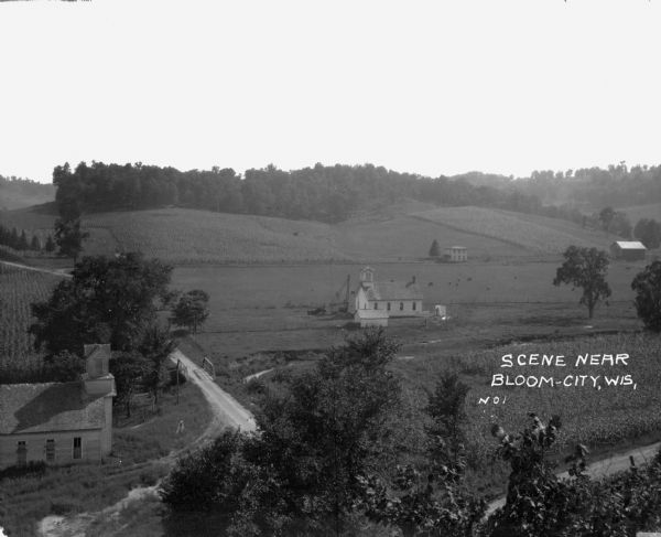 Elevated view of farmland. In the bottom-left center, there is a structure with a bell tower near a dirt road. In the center there is a similar building. There is a tractor and machinery in the yard. Cattle graze in a field in the background near what may be a farmhouse.