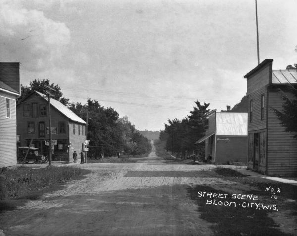 View down a long dirt road toward an intersection with two shops. On the left is a car parked outside the shop. Three men pose together at the corner. There are signs posted with the words, "August 21." On the right a man sits on the stoop of the shop with his dog.