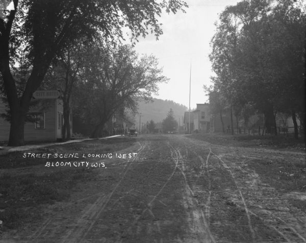 View down a dirt road. The City Hotel is on the left. Farther down the road on a sidewalk a man talks on a platform while a group of men listen.