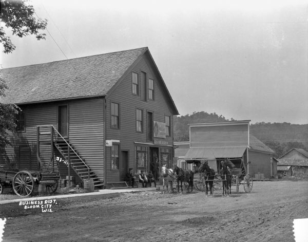 A street scene at the post office. The post office houses J.E. James Childs Cabinets. One man stands at the doorpost and three men sit on makeshift seats on the sidewalk in front of the post office. Four horses and two carriages stand at the street.
