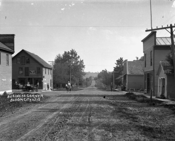 View down dirt road of an intersection with a man on a horse standing in the middle. On the left there is Bradley & Metcalf C.(?) Bradley shoe store. Men stand in front of the store. A car, with two men inside, is parked at the entrance.