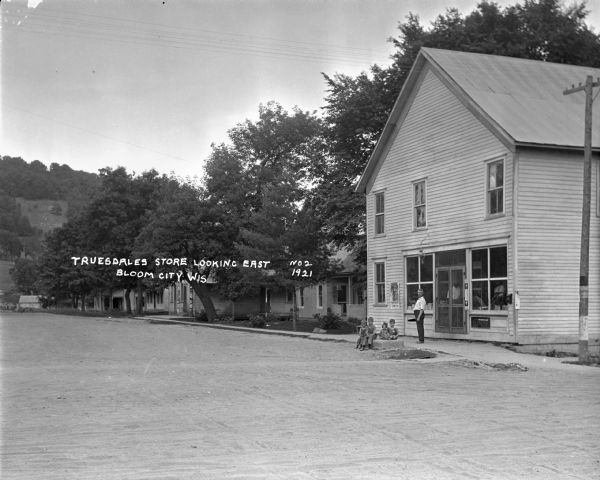 View from street of exterior of the Truesdale's store where a man and four children pose on the sidewalk. There are homes and a barbershop pole [?] on the street further down.