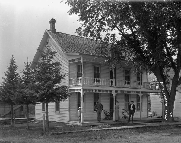 Exterior of a two-story hotel with two men, a woman and a child standing on the porch.