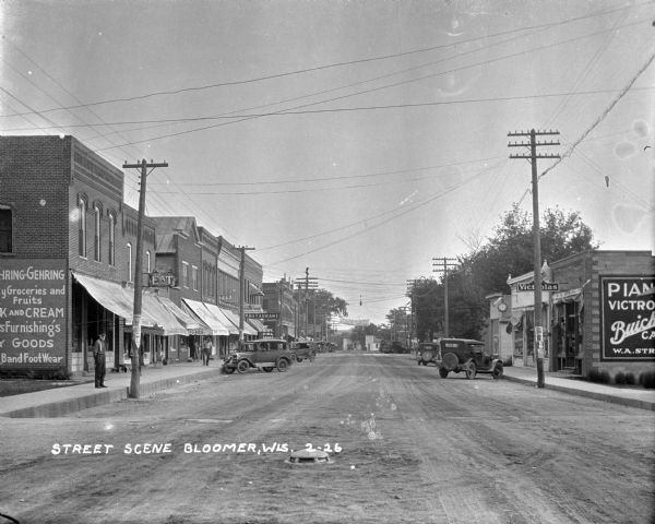 View down retail street. Shops and parked cars are on either side. On the side of a shop on the left there is a hand-painted advertisement for the selection at a general store. The visible storefronts are a general store, cigar shop, restaurant, drug store, and S.A. Struve Victor-Victoria store and Buick car dealership. There is a banner going across the road advertising a pageant.