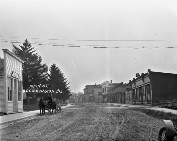 A man driving a carriage pulled by two horses is at the curb of the post office. On the street, there is the dentist office of H.H. Hancock, a bank, and J.D. Ludden general merchandise. A utility line hangs across the frame. The tire of a car is in the right corner.