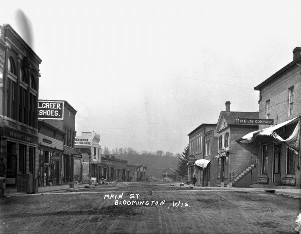 Shops along Main Street. On the left, there is a Woodhouse and Bartley bank, a hardware store, Greer shoe store, J.B. Ludden general merchandise, a bank, and H.H. Hancock dentist. On the right, There is R.E. Jay Company clothing store, Ora Hatch Druggist drug store, and Wall Paper and Paint store. A man stands in the doorway at the hardware store and another man walks on the sidewalk near the drugstore.