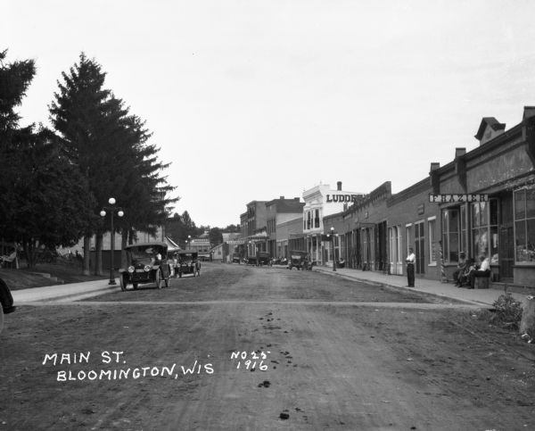 View of Main Street from the middle of the street looking down the street. Cars are parked on either side of the street. On the left side, there is a woman sitting on a chair on the lawn and reading a book, and a cafe. The shops on the right are: "Frazier," a bank, J.B. Ludden general merchandise, and Ludden's men's clothing store. Four men are seated and one man stands in front of Frazier. There is horse dung in the street.