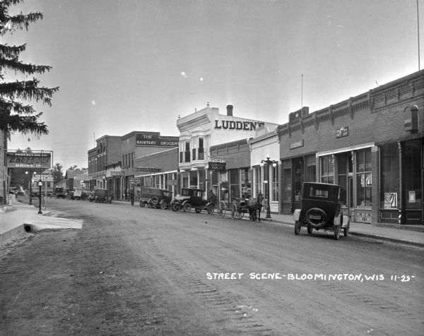 View down middle of Main Street. The right side of the street features: Bloomington State Bank, Ira Fox Meat Market, C.H. Enke, Sprague's Recreation Parlor, Oates and Ellis Hatchery, Ludden's Store for Men, Mulrooney Restaurant and Drug Department, Groceries and Meats, Woodhouse and Bartley Bank, Clem-Meyer Dry Goods and Groceries. Perfect Oil Company Energee! Drive in is on the left side of the street. Cars, a horse and carriage, and a milk truck are parked at the curb.