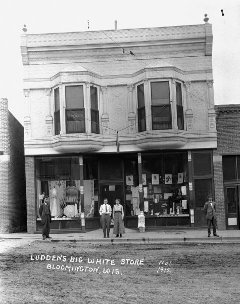 The facade of Ludden's Big White Store. The building features two bay windows, two stories, and two large display windows. The window displays umbrellas, dresses and hats. Three men and a woman stand on the curb.