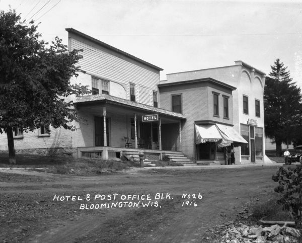 A hotel, barbershop [?], and post office. The hotel has a roofed porch and chairs. A man stands under the awning of the barbershop, and another man leans against a car on the far right.