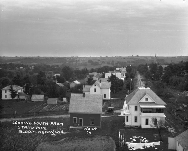 Elevated view from the water tower. There is a laundry line in the backyard of the white house in the foreground. A car is driving down the road. In the distance, there is a church steeple, barns and farmland.