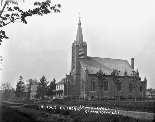 The exterior of the Catholic church and the parsonage. The church building has a gable roof, lancet arched windows, and a steeple with a cross atop.