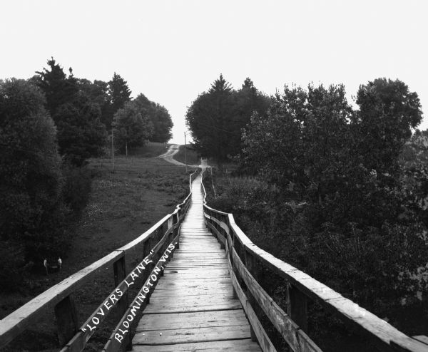 View across a wooden footbridge. There are two men and a cart below on the left.