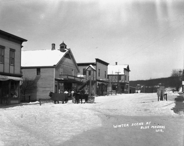 A snow-covered street with several horses standing at the curb. Groups of people pose on the porches of the hotel and the post office.